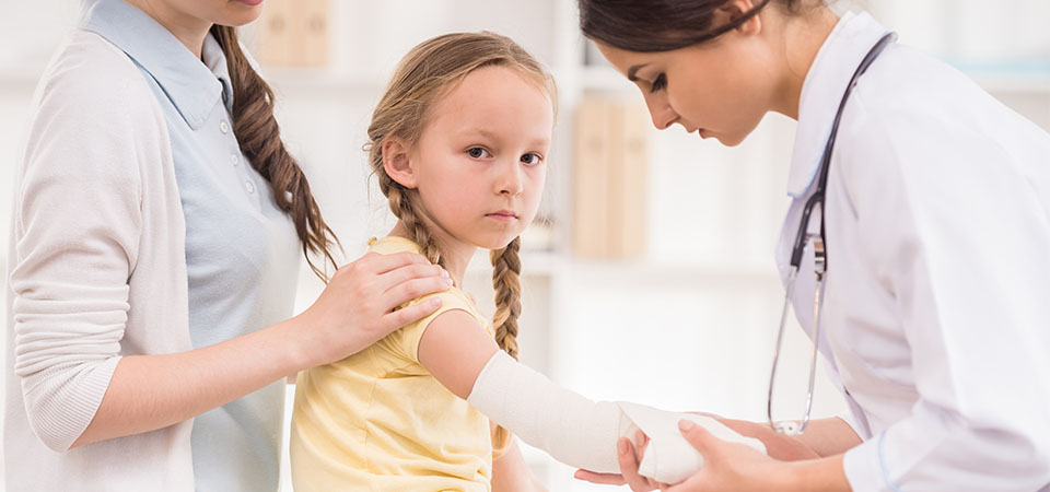 En læge undersøger en piges arm, mens en voksen holder hende. En medicinsk person tilser hendes sår.